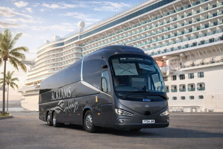 A black shuttle bus with the words 'ACKLAMS' and 'Shuttles' on its side parked on a street near a large cruise ship docked at a port, with palm trees nearby and a partly cloudy sky above.