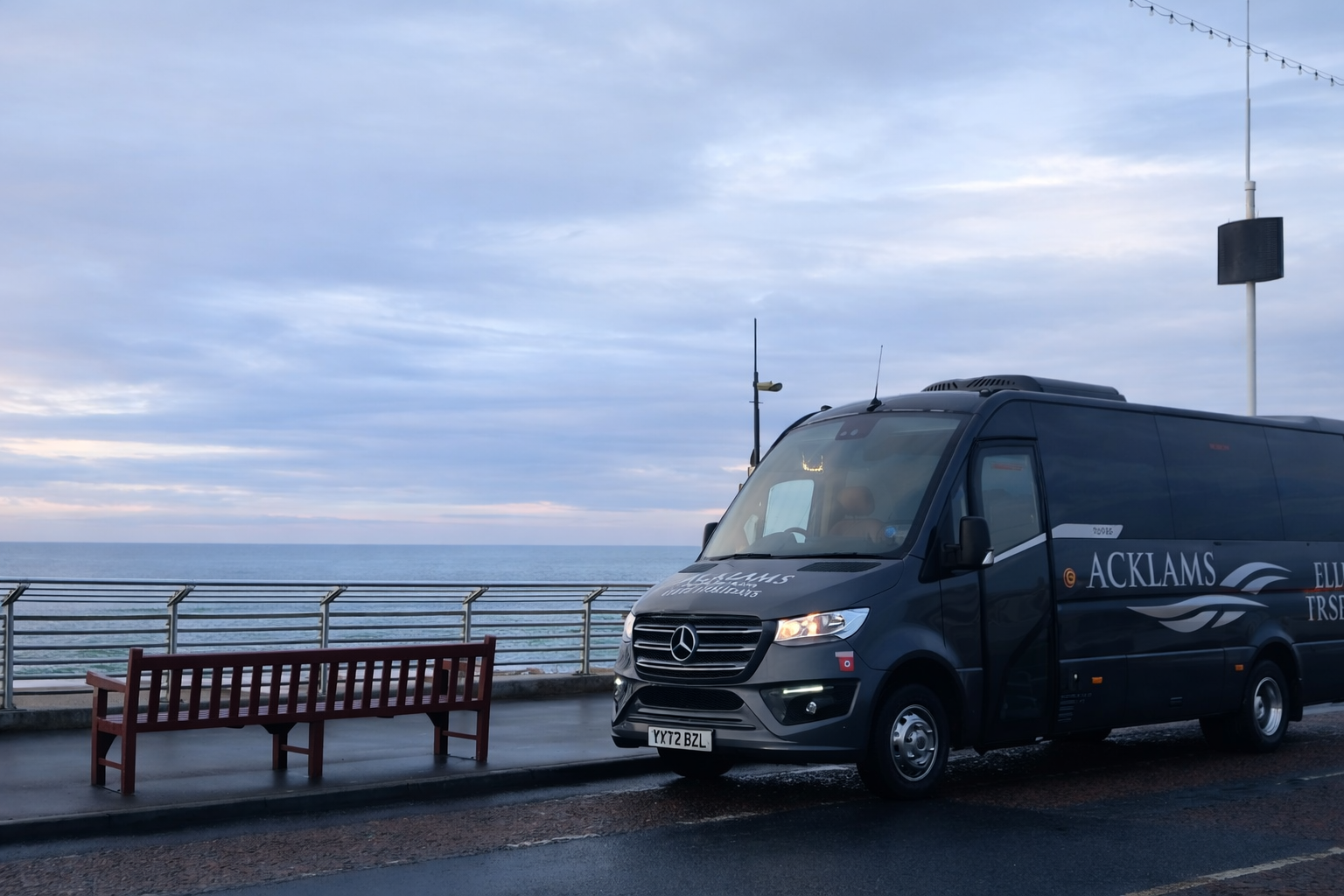A black Mercedes-Benz van parked near a seaside promenade with a red bench and metal railing, overlooking the ocean on a cloudy day.