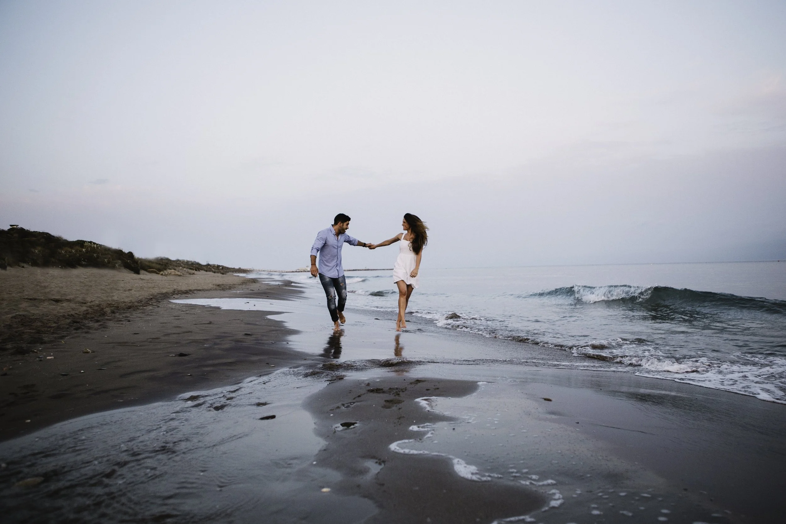 A couple is walking hand in hand on a beach, near the shoreline with waves, under a cloudy sky.