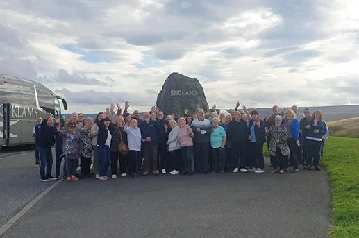 Group of people standing in front of a large rock with 'England' engraved on it, near a bus and an ocean view under a cloudy sky.