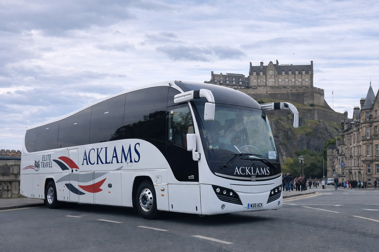 A white coach bus with black accents labeled 'ACKLAMS' and 'ELITE TRAVEL' parked on a city street with Edinburgh Castle visible in the background and a group of people walking on the sidewalk.