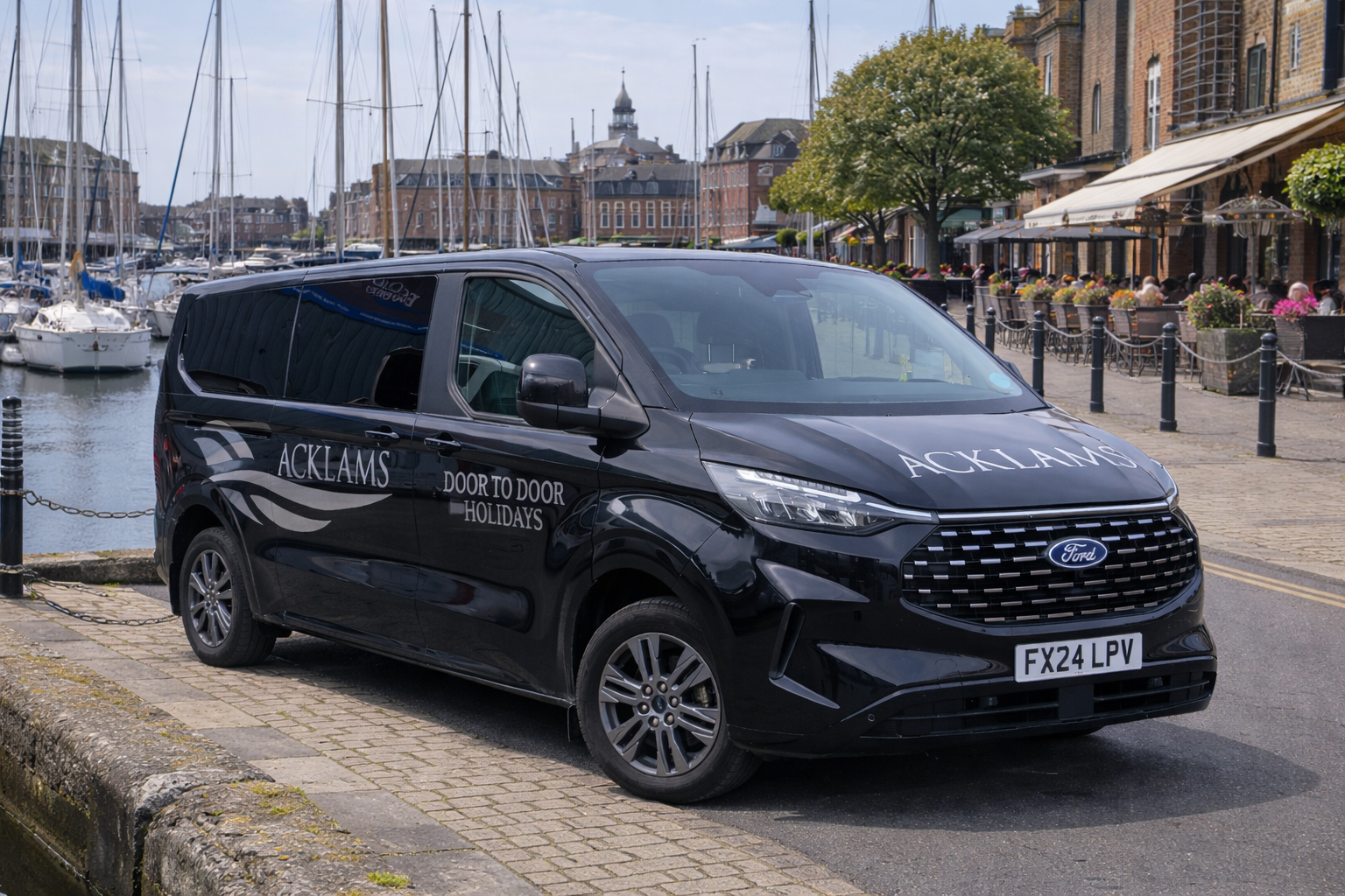 Black Ford van parked along a waterfront in a town with boats docked in the harbor and outdoor seating at cafes and restaurants with people dining, on a cloudy day.