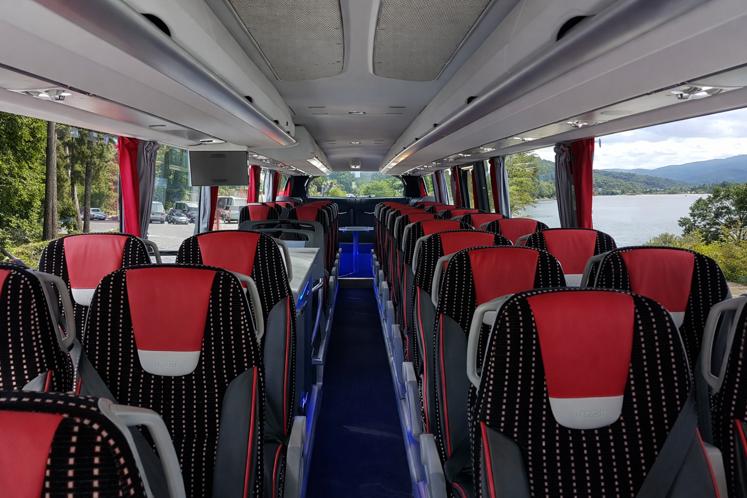 Interior of a tour bus with red and black seats, facing forward, with large windows showing a lakeside landscape of water, trees, and mountains.