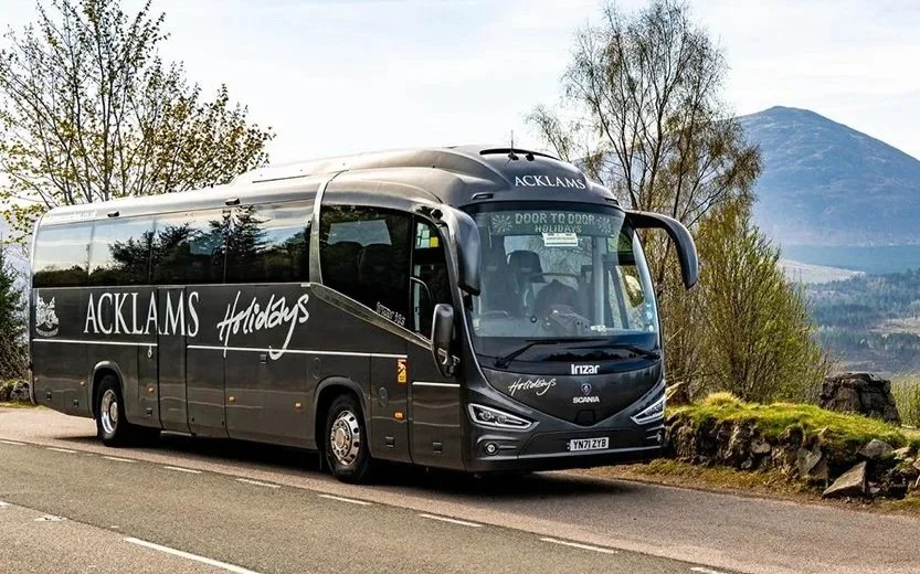 A black Acklams Holidays tour bus parked on the side of a road, with trees and mountains in the background.