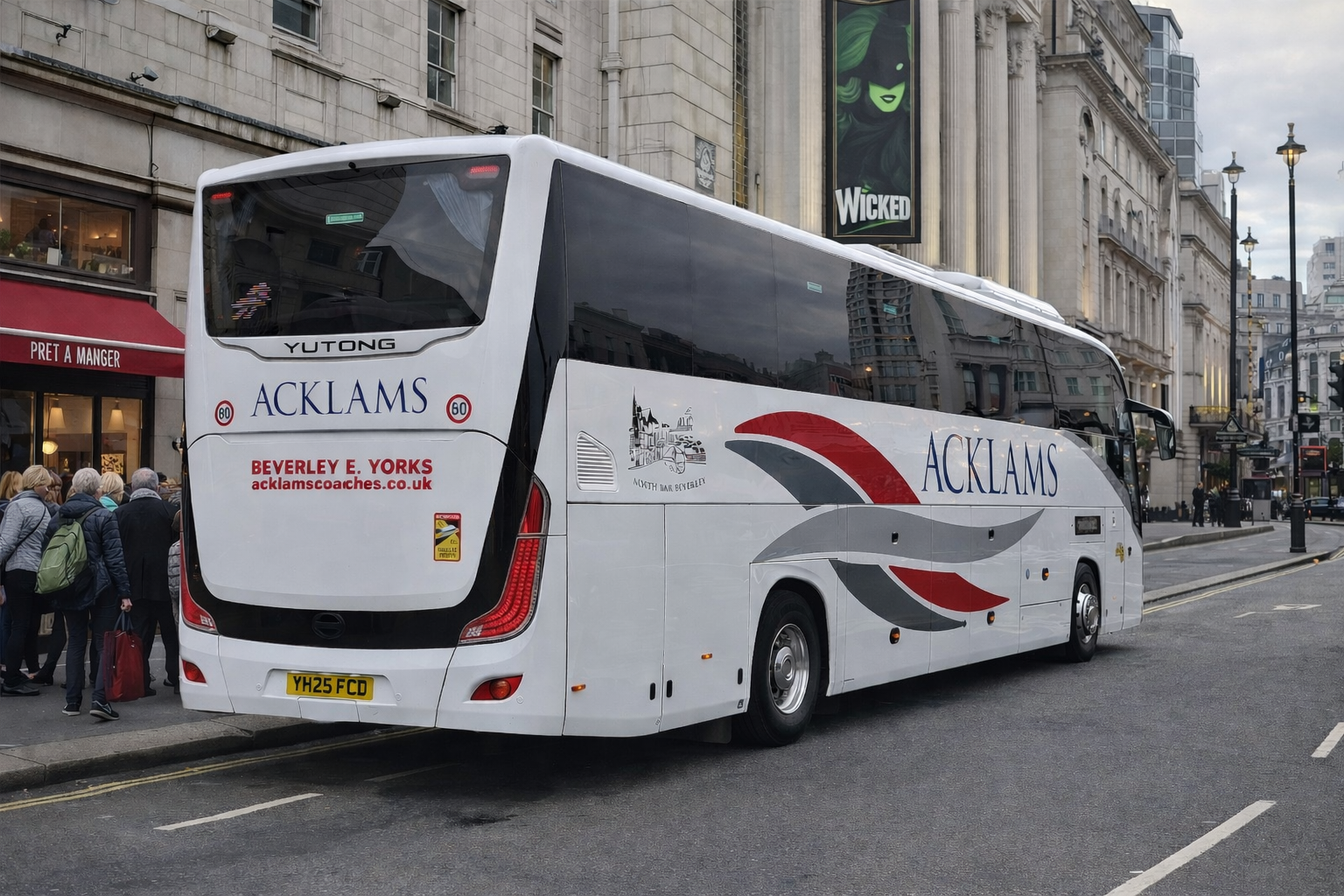 White coach bus with 'Acklams' branding parked on city street, with people walking and a theatre billboard advertising 'Wicked' in the background.
