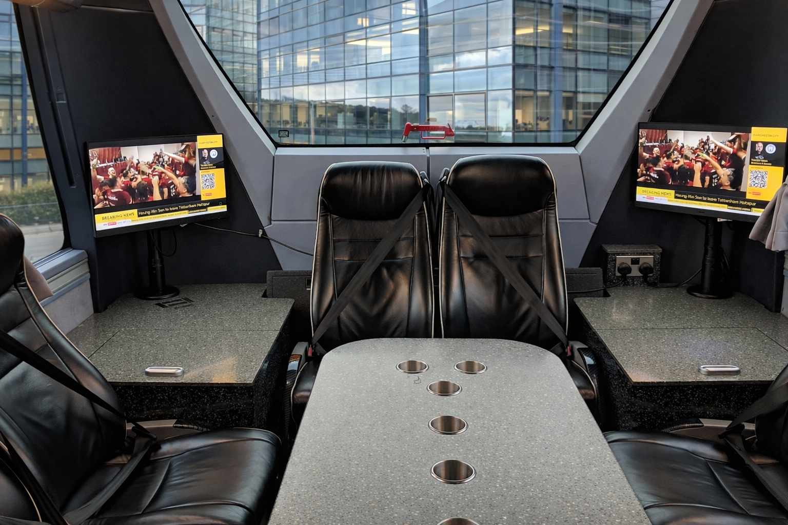 Indoor meeting room with black leather chairs, two computer screens displaying a meeting, a table with built-in cup holders, and large windows showing an office building outside.