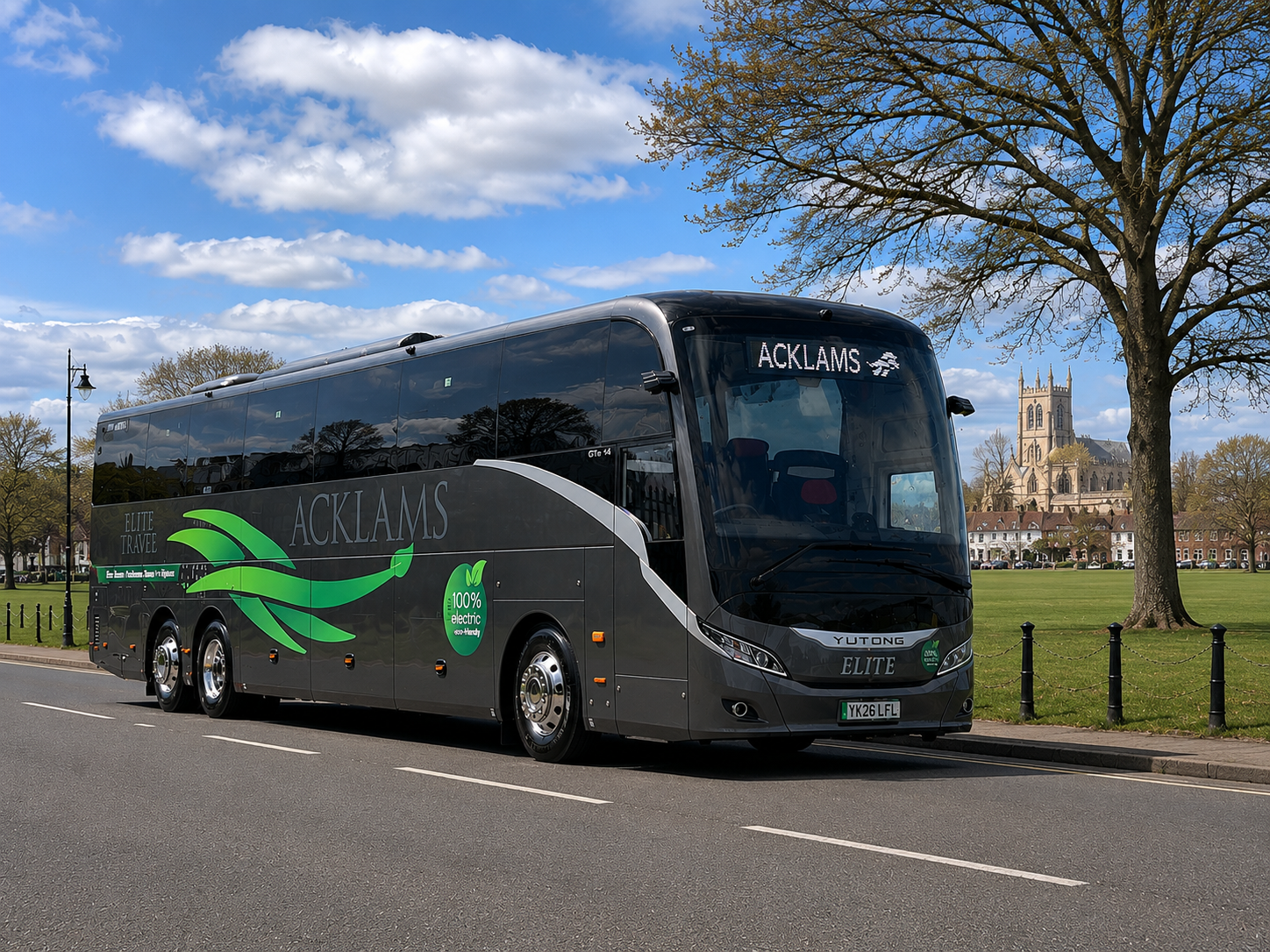 Grey electric coach bus with green graphics and 'ACKLAMS' branding driving on a city street near a park with trees and a historic church in the background.