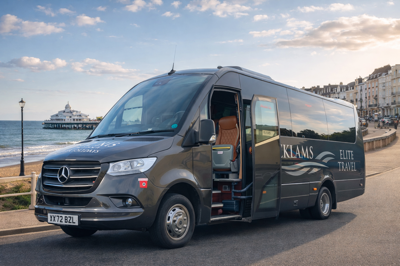Black Mercedes-Benz shuttle bus parked on street by beach, with pier, ocean, and residential buildings in background.