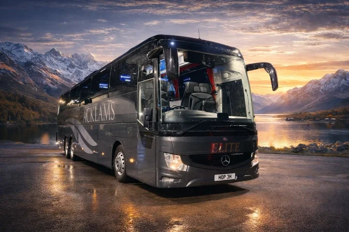 Black luxury bus with 'ACKLANDS' branding parked near a lake with mountains in the background during sunset.