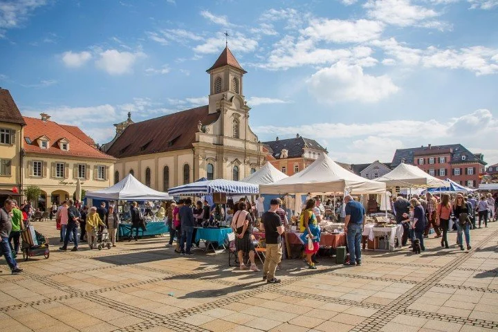 Outdoor market with tents set up in a town square, people shopping, and a church with a steeple in the background.