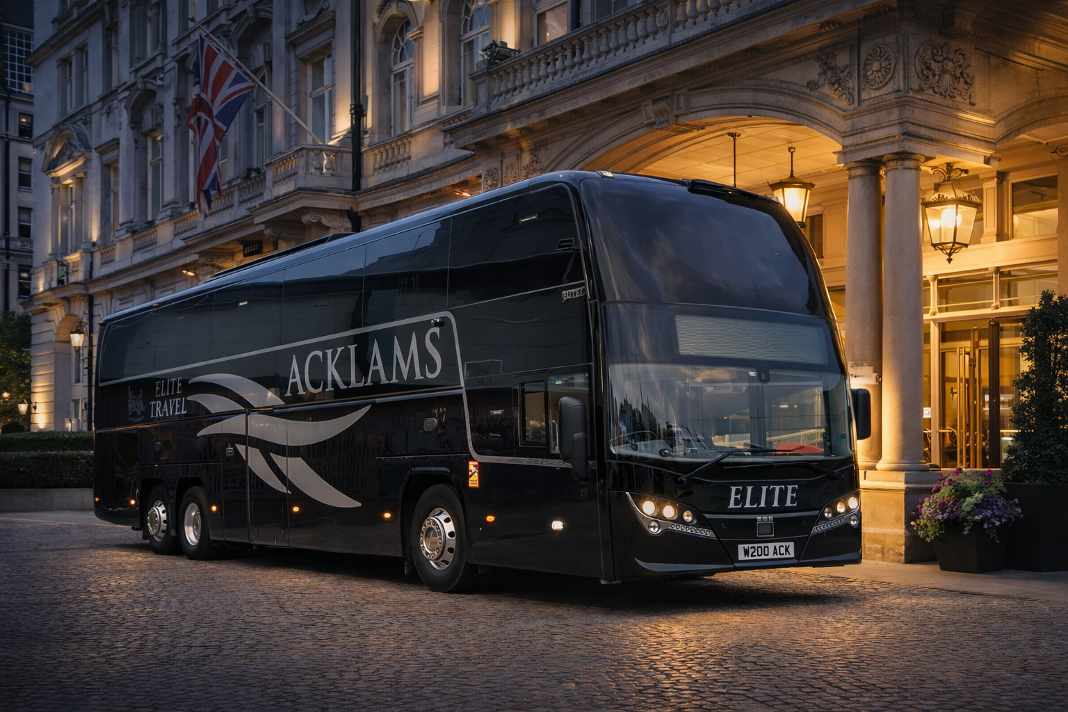 A black luxury bus with the branding 'ACKLAMS ELITE' parked outside an elegant building with columns and warm lighting at dusk.