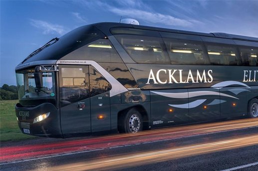 Black luxury Coach bus with 'ACKLAMS' branding, parked on a road at sunset.