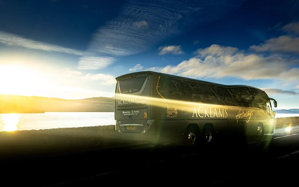 A black tour bus with the words 'ACKLAMS Holidays' written on the side, parked beside a body of water during sunset or sunrise, with clouds in the sky.