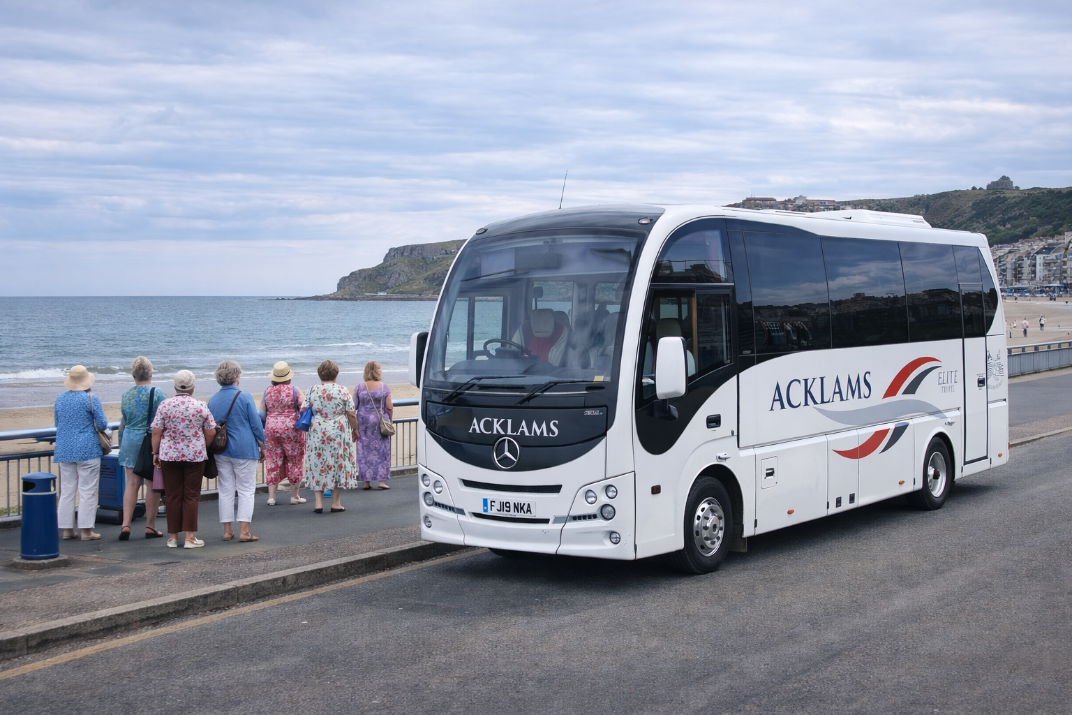 A group of elderly women in colorful dresses and hats standing on a seaside promenade, looking at the ocean. A white tour bus with "ACKLAMS" branding is parked nearby, with a coastal town and cliffs in the background.