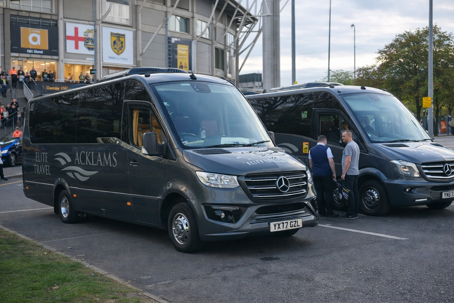 Two black Mercedes-Benz vans labeled 'Elite Travel' and 'Acklams' parked outside a stadium with people and flags in the background. Two men are standing beside the second van, talking.