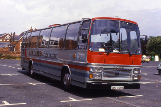 A vintage passenger bus parked in a parking lot, with buildings and trees in the background.