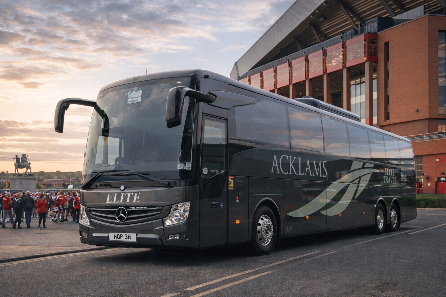 A black luxury coach bus with Mercedes-Benz logo parked on a city street, with a crowd of people and a monument featuring a mounted rider statue in the background, and a modern building with glass and brick facade at sunset.