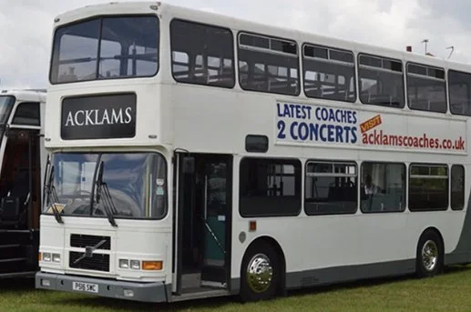 A white double-decker bus parked outdoors with advertisements for Acklams Coaches and upcoming concerts.