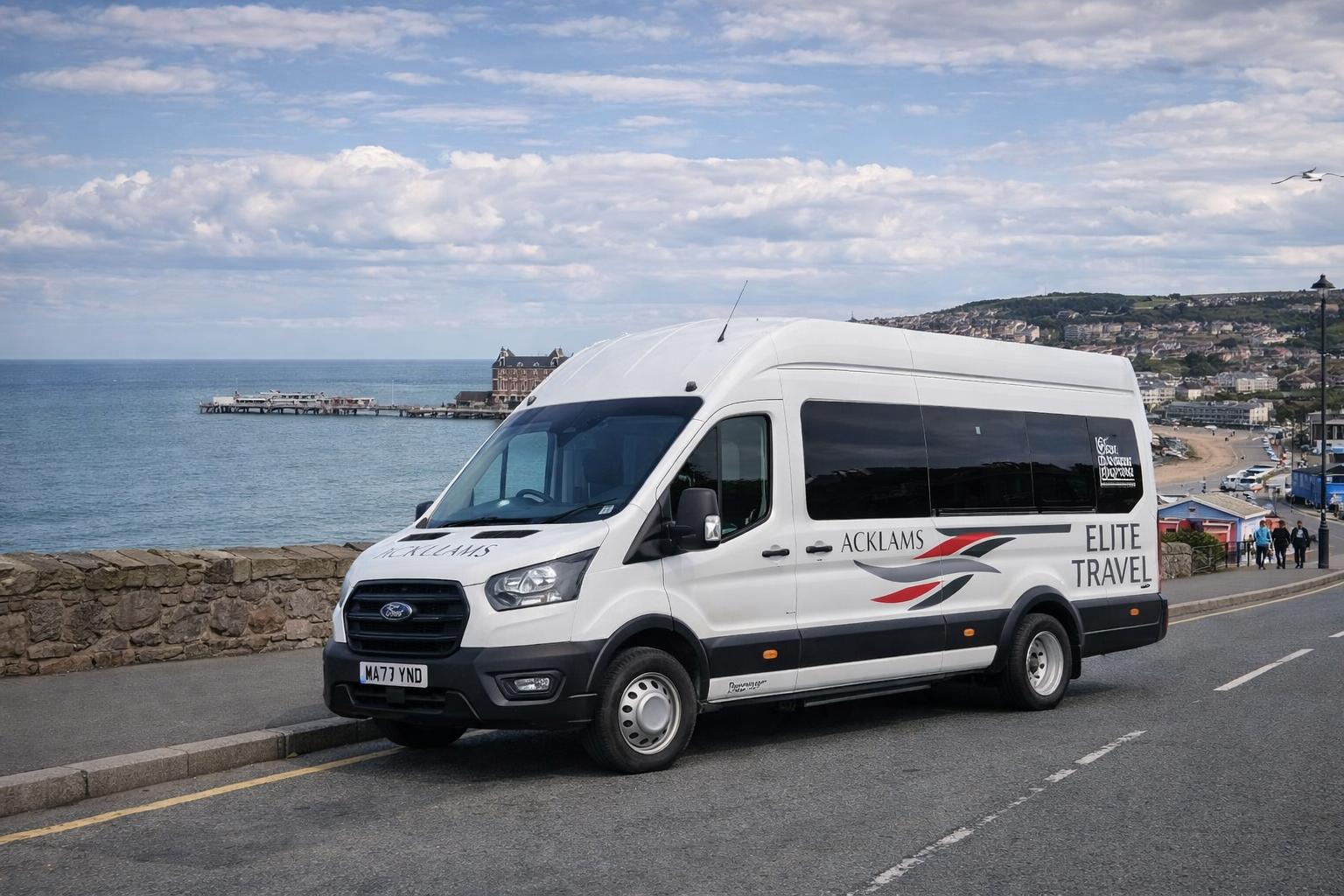 White Ford transit van with 'ACKLAMS ELITE TRAVEL' lettering parked by a stone wall overlooking the ocean, with a pier and buildings in the background on a partly cloudy day.