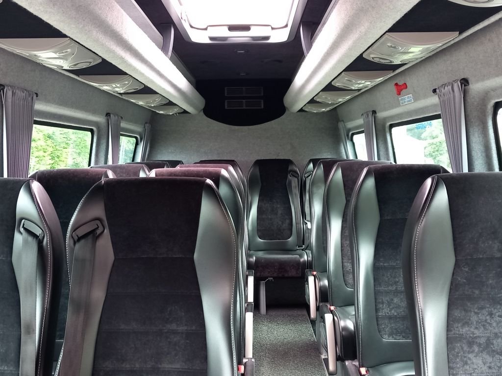 Interior of a bus with multiple black and gray seats, windows with gray curtains, overhead compartments, and a ceiling with a large white panel.