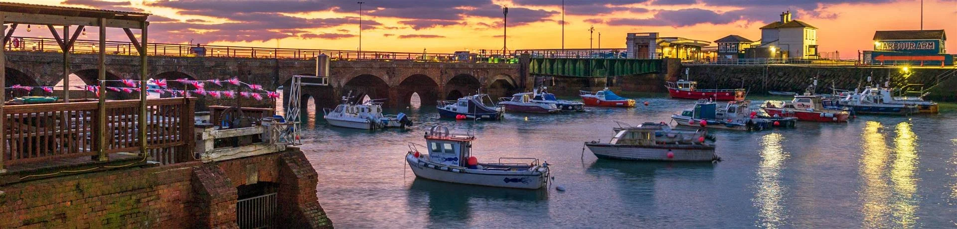 Boats docked in a harbor at sunset with a brick pier and a bridge in the background.