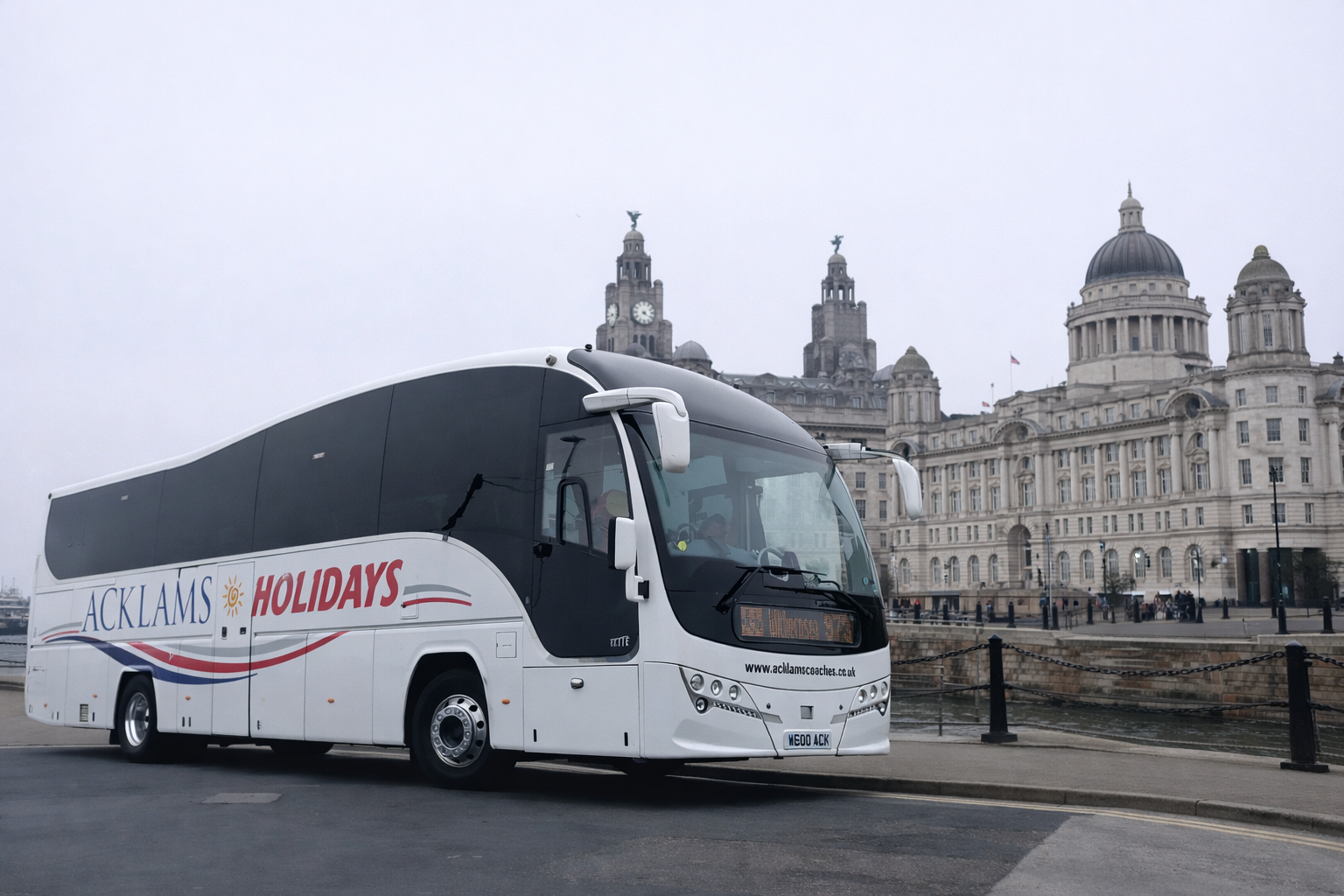 White coach bus with 'ACKLAMS HOLIDAYS' branding parked near a river with historic buildings in the background.