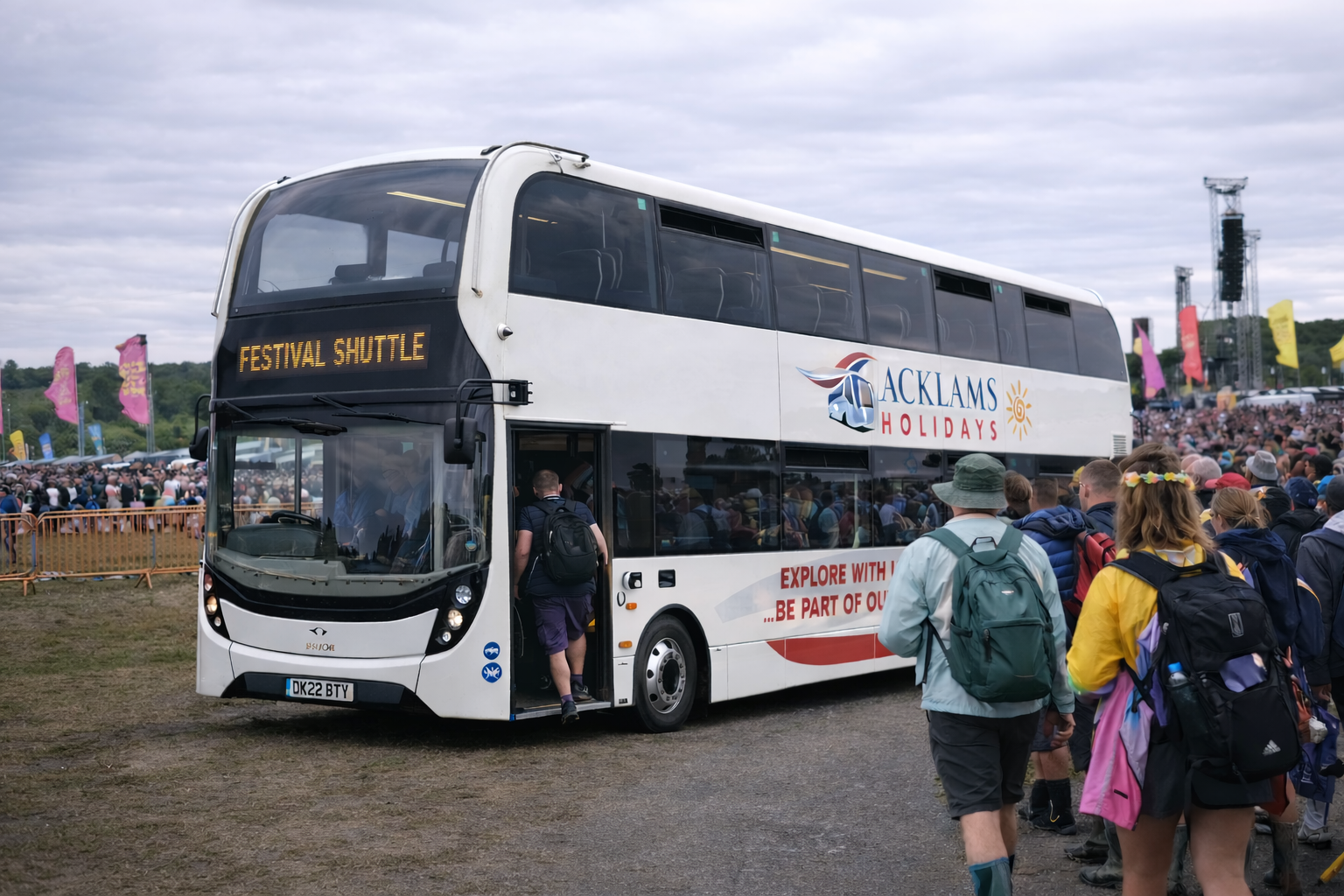 Outdoor festival scene with a crowd and a double-decker shuttle bus labeled 'ACKLAMS HOLIDAYS' and 'FESTIVAL SHUTTLE'