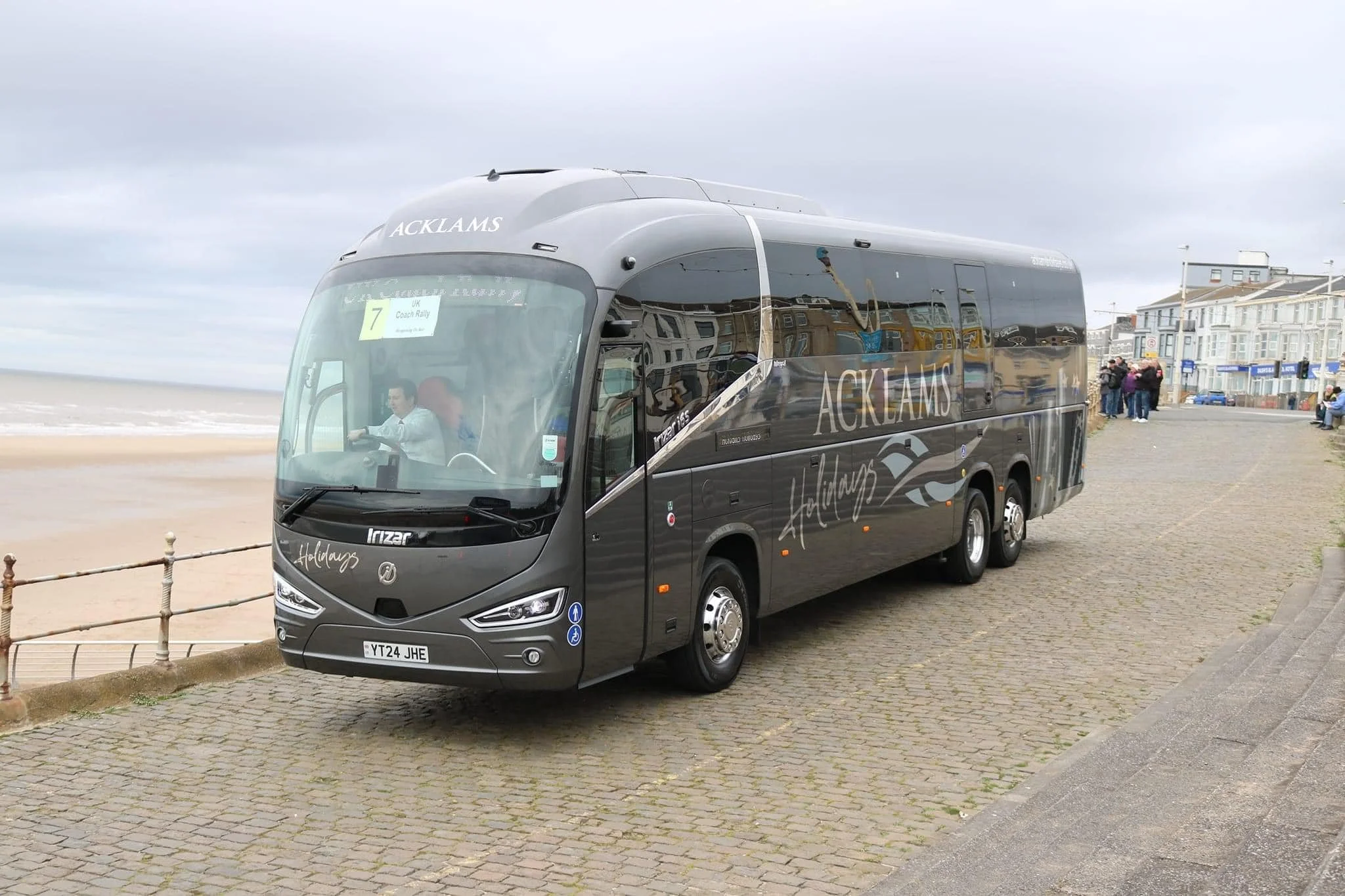 A black Acklams Holidays bus parked on a cobblestone promenade near the beach, with a group of people and colorful buildings in the background.