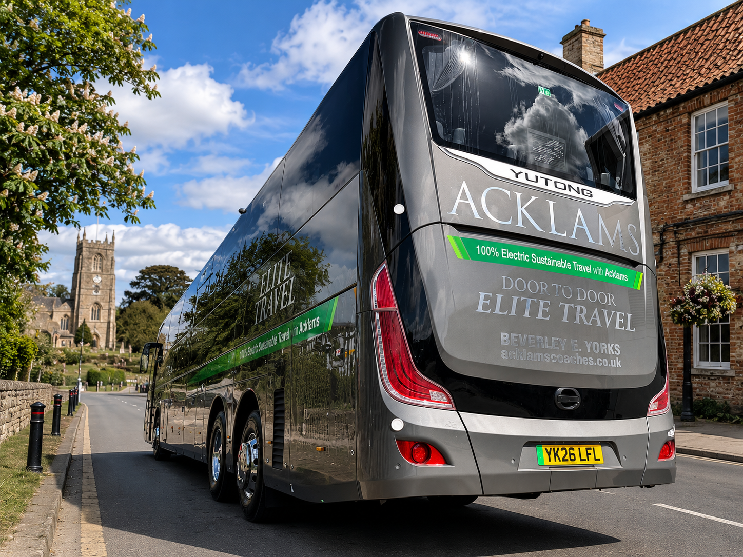 A sleek black double-decker bus parked on a street with a historic church and brick buildings in the background. The bus has signage indicating it is a 100% electric vehicle operated by Acklams for Elite Travel, with a license plate YK26 LFL.