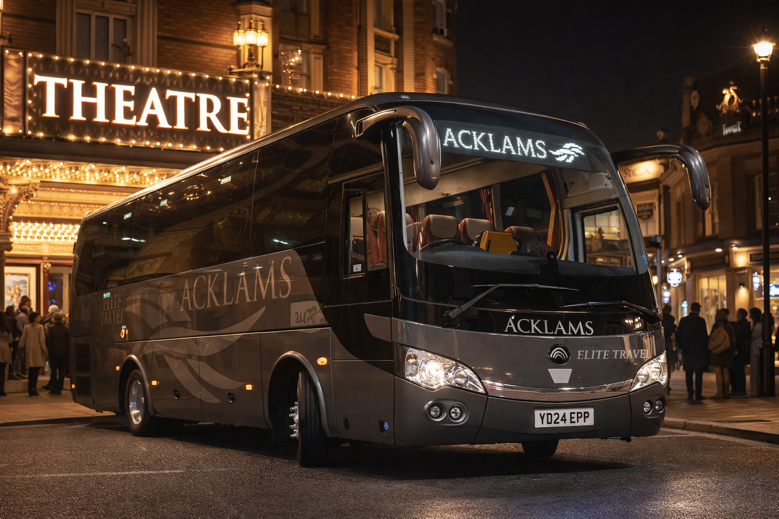A black bus with 'ACKLAMS' and 'ELITE TRAVEL' written on it parked on a city street at night, with a theater marquee and pedestrians in the background.