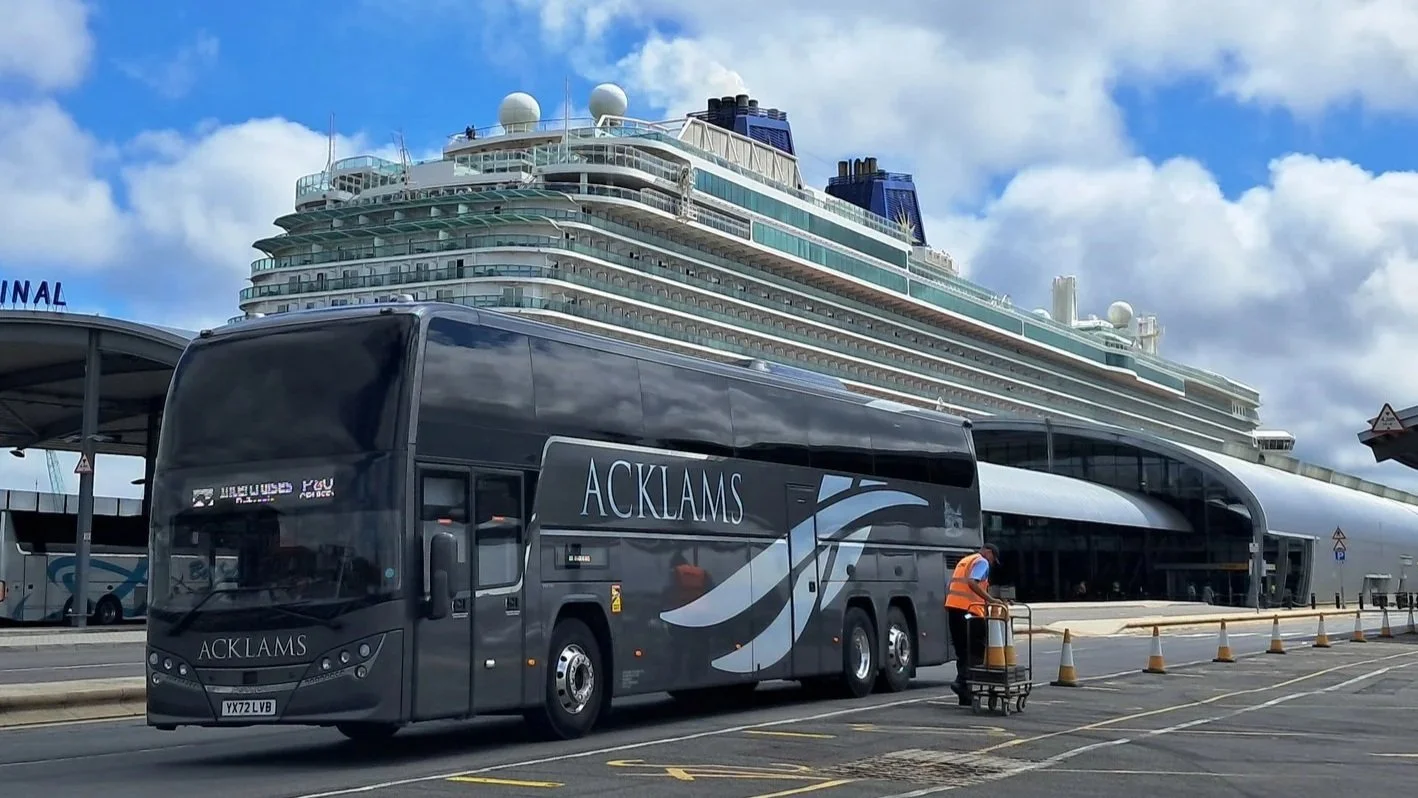 A large cruise ship docked at a port with a black bus labeled 'ACKLAMS' parked in front. A worker in an orange safety vest is pushing a trolley near the bus, with orange traffic cones outlining the area. The sky is cloudy.