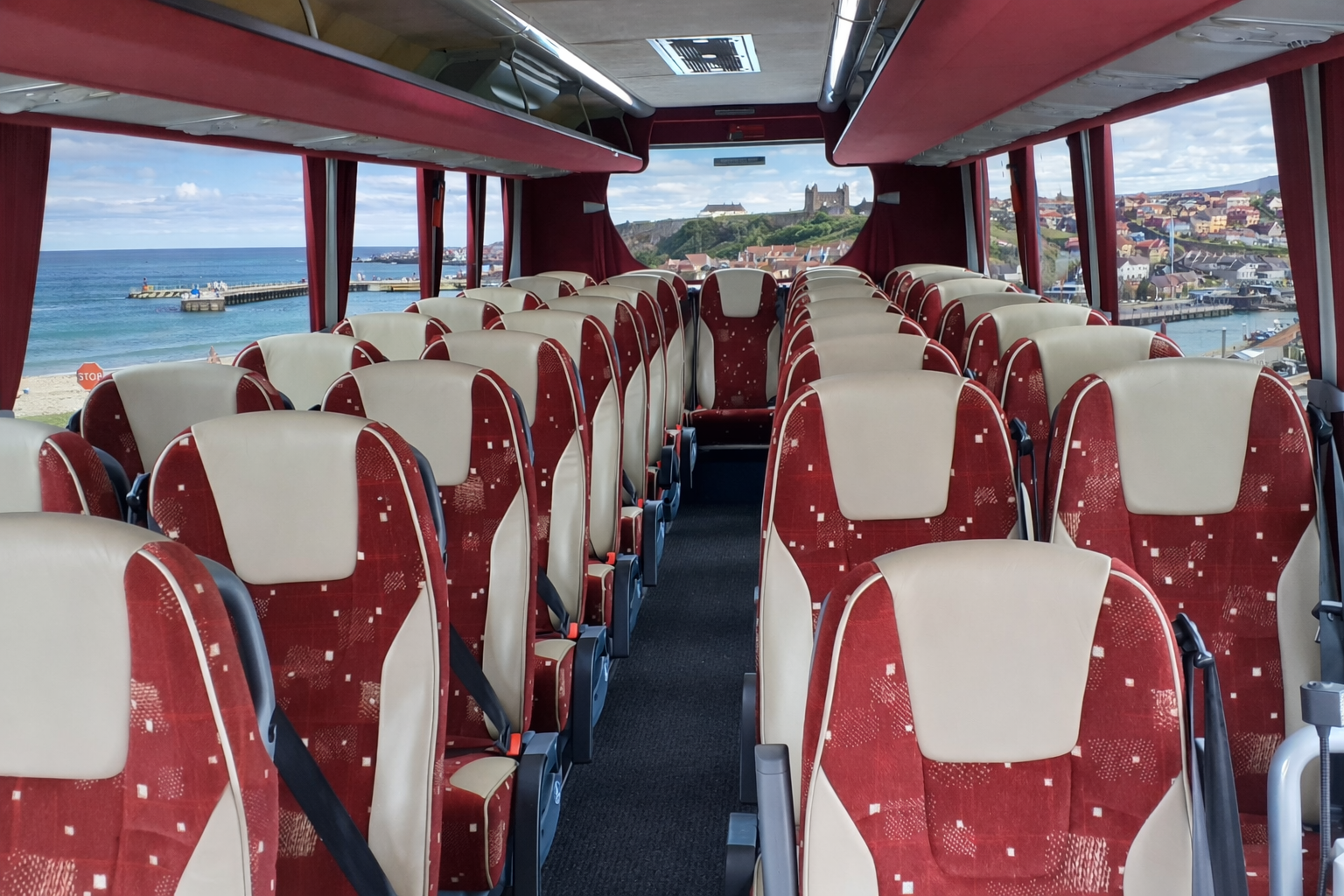 Inside a coach bus with red and beige seats, large windows showing a seaside town with houses, a pier, and a castle on a hill in the background.