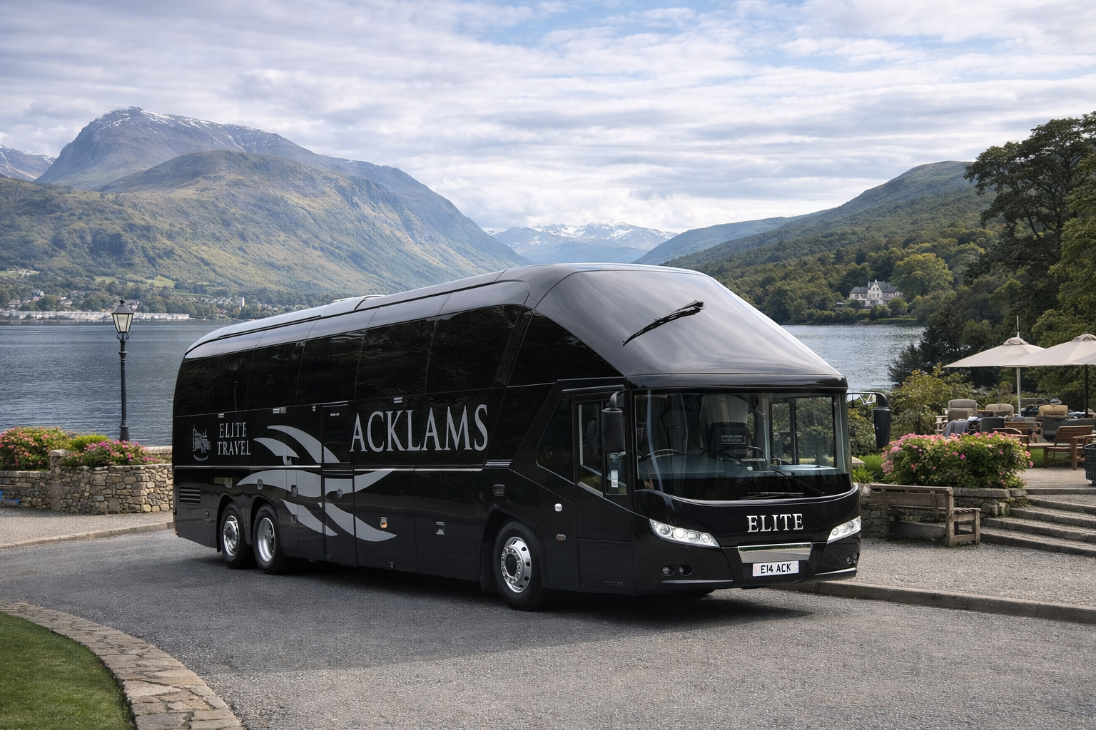 A black luxury tour bus labeled 'ELITE' and 'ACKLAMS' parked near a lakeside with mountains and greenery in the background, with outdoor seating and umbrellas nearby.
