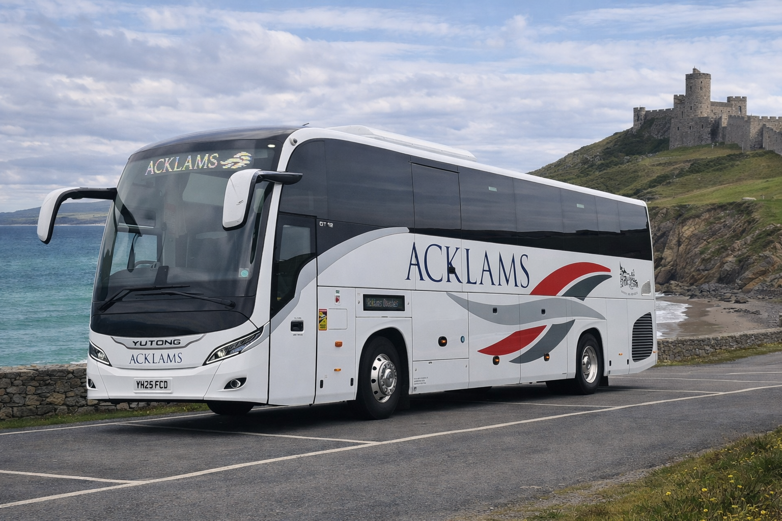 A white Acklams bus parked on a roadside near a beach, with a castle on a hill in the background under a cloudy sky.