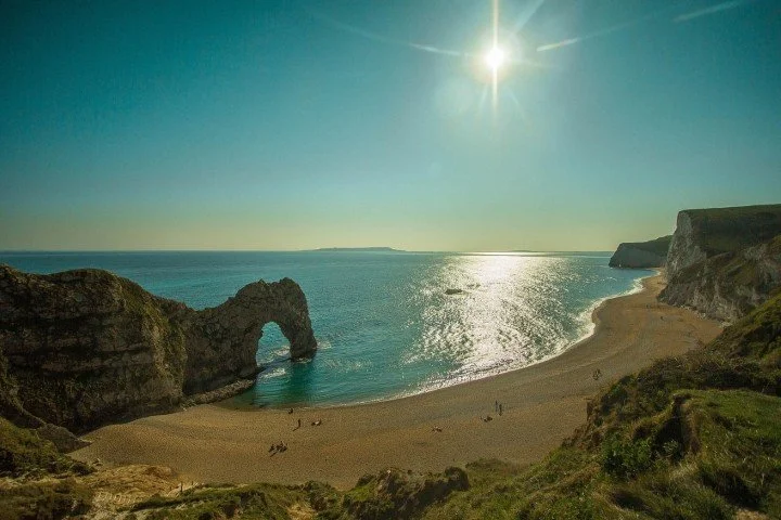 Scenic view of a beach with a natural rock arch, ocean waves, and cliffs under a bright sun.