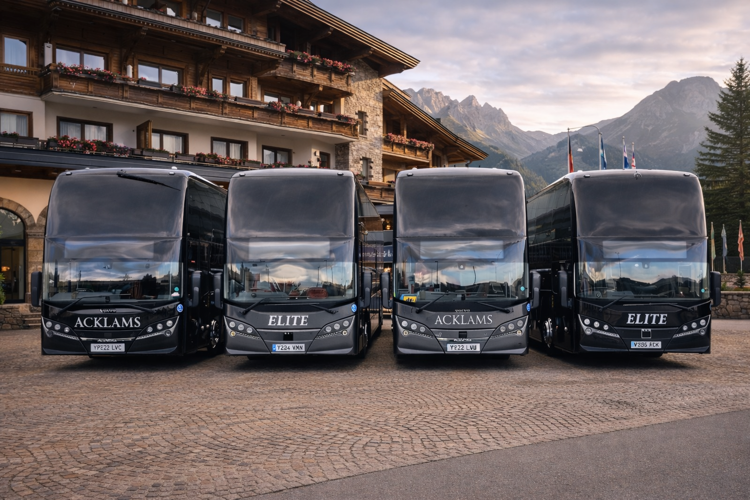 Four black touring buses parked in front of a multi-story wooden hotel with balconies and flower boxes. Mountain peaks are visible in the background during cloudy weather.