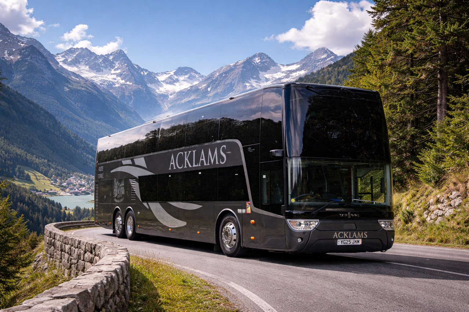 A large black bus with the word "ACKLAMS" on the side, traveling along a mountain road with a stone guardrail. The background features snow-capped mountains, green forests, a blue sky with white clouds, and a small village near a lake.
