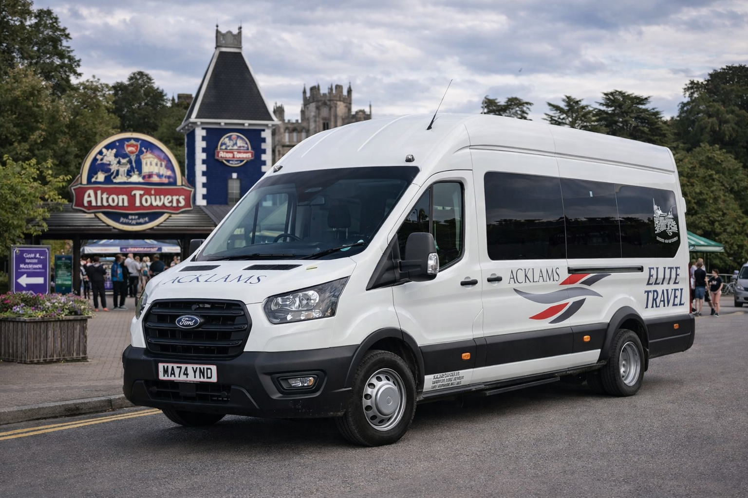 White Ford transit van with 'Acklams Elite Travel' logo parked near Alton Towers entrance with people and castle in the background.