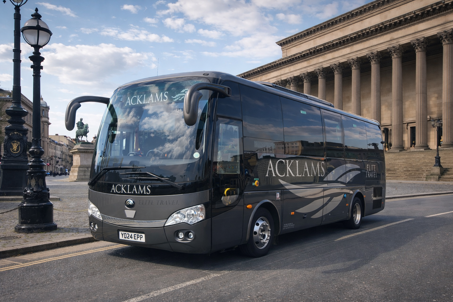 A black coach bus with the words ACKLAMS and ELITE TRAVEL on the side, parked on a city street in front of a historic building with columns, lamp posts, and a statue of a man on horseback in the background.