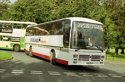 A white bus with red stripe and 'ACKLAMS' written on front and side, parked on a road with green trees in the background, and another bus visible behind it.