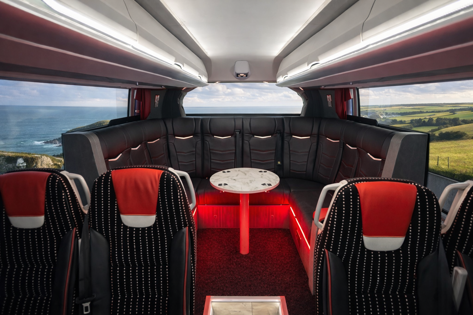 Inside a tour bus or coach with black leather seating arranged in a U-shape, a small marble-topped table in the center, large windows showing a scenic coastal landscape, and overhead storage compartments.