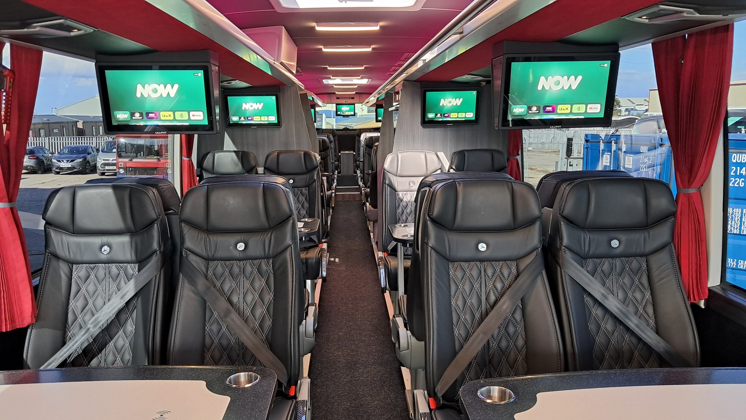 Interior of a modern bus with black leather seats, multiple overhead screens displaying 'NOW,' operating curtains on windows, and a view of parked cars and blue recycling bins outside.