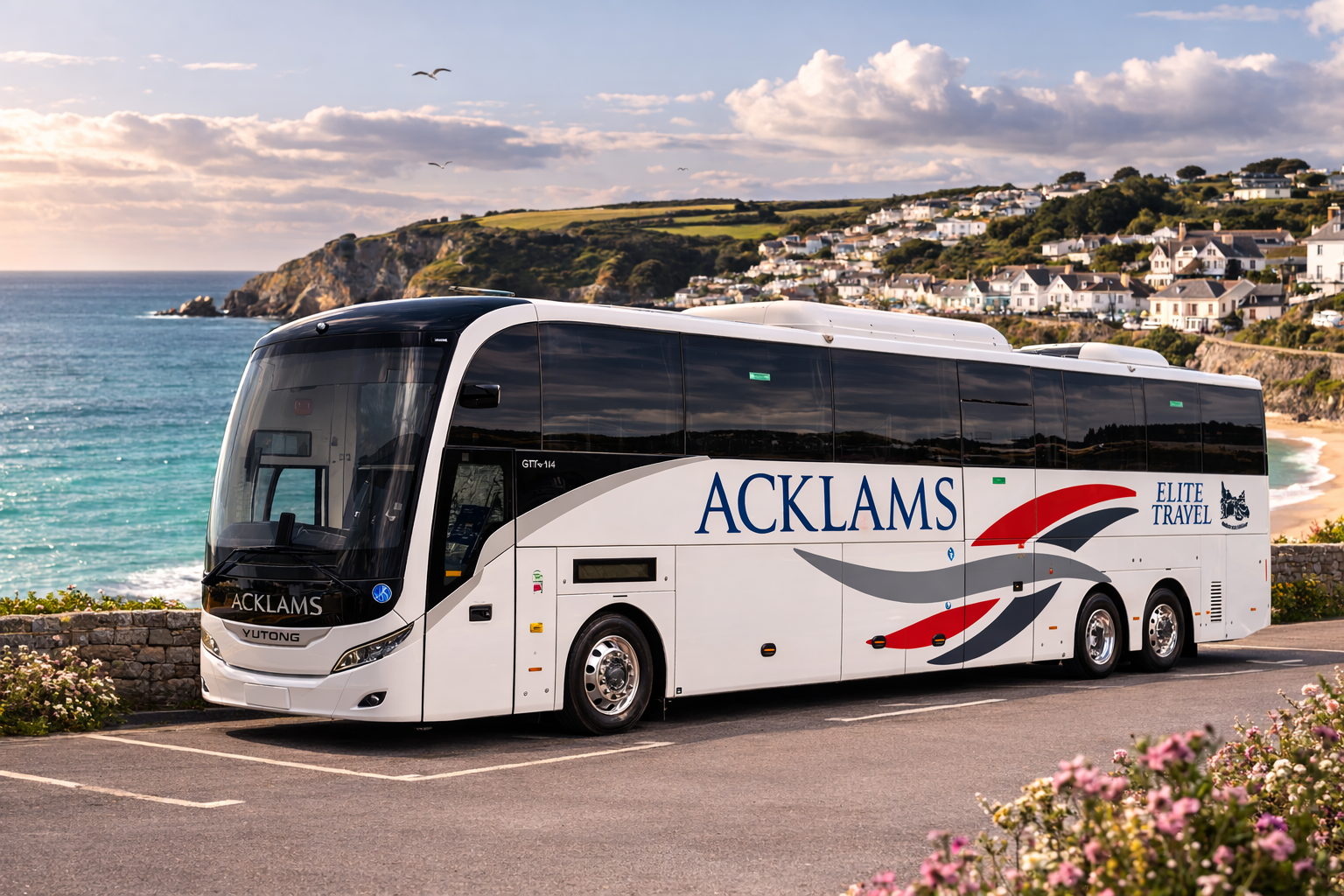 A white Acklams tour bus parked near the coast with a background of a rocky shoreline, ocean, and a hillside residential area under a partly cloudy sky.