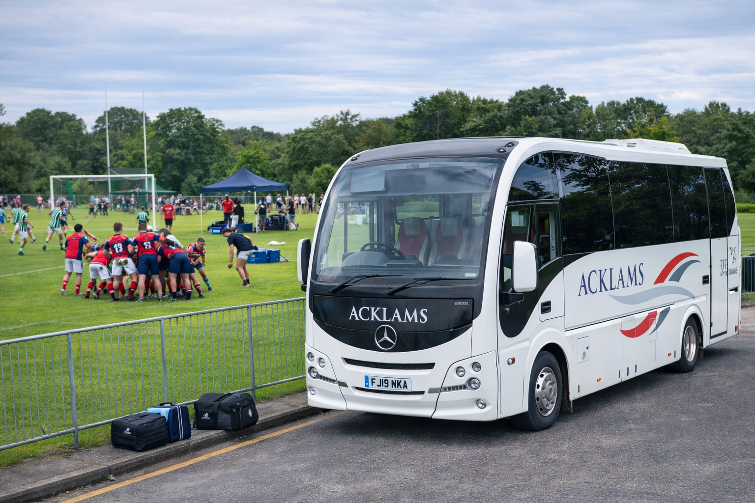 A white Acklams bus parked near a rugby field where players in red and navy uniforms are engaged in a scrum during a game. There are spectators, tents, and trees in the background.