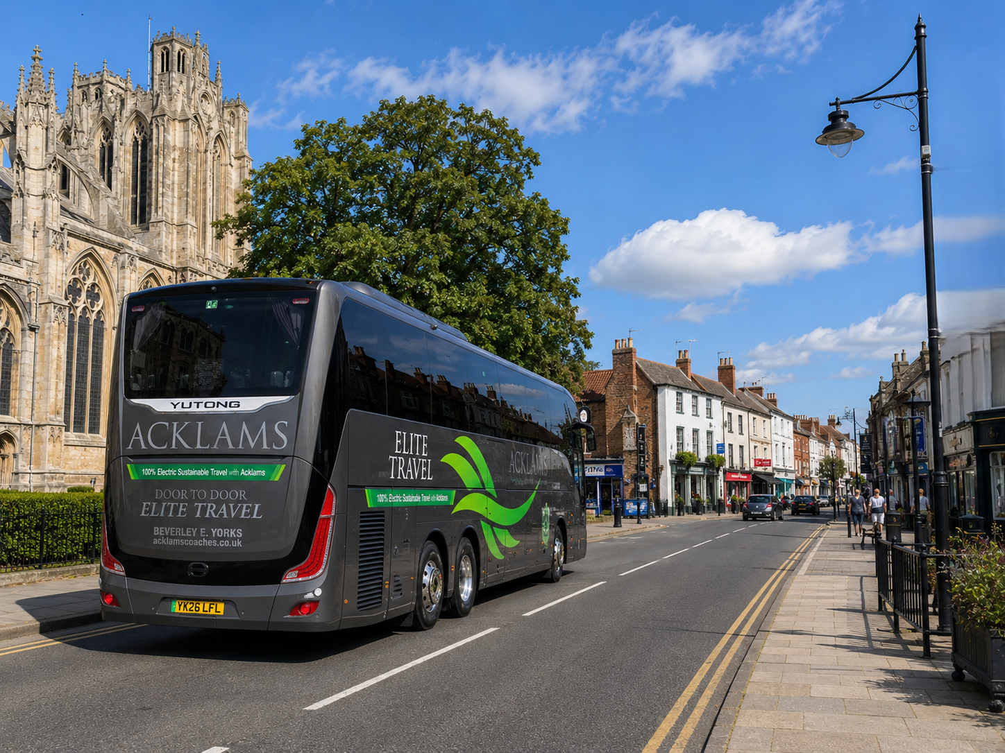 A  grey electric bus labeled 'Elite Travel' on a city street with historic buildings, a church, and pedestrians under a partly cloudy sky.