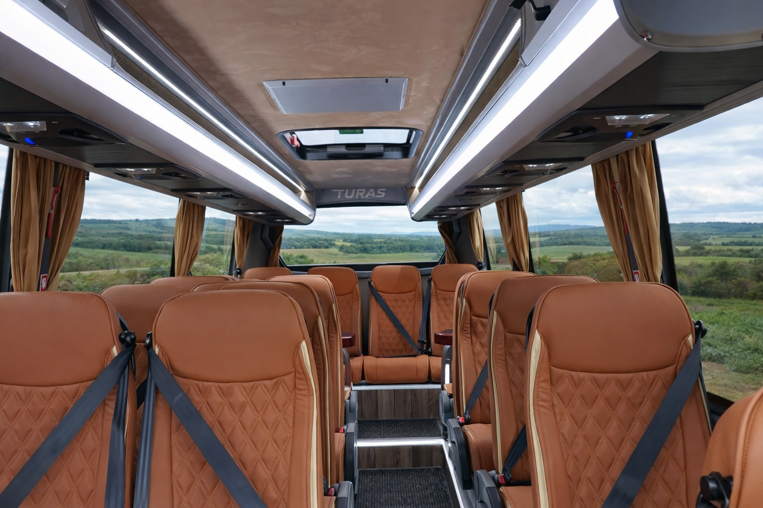 Inside a modern bus with tan leather seats, large windows, and scenic countryside view, featuring overhead lighting and curtains on the windows.
