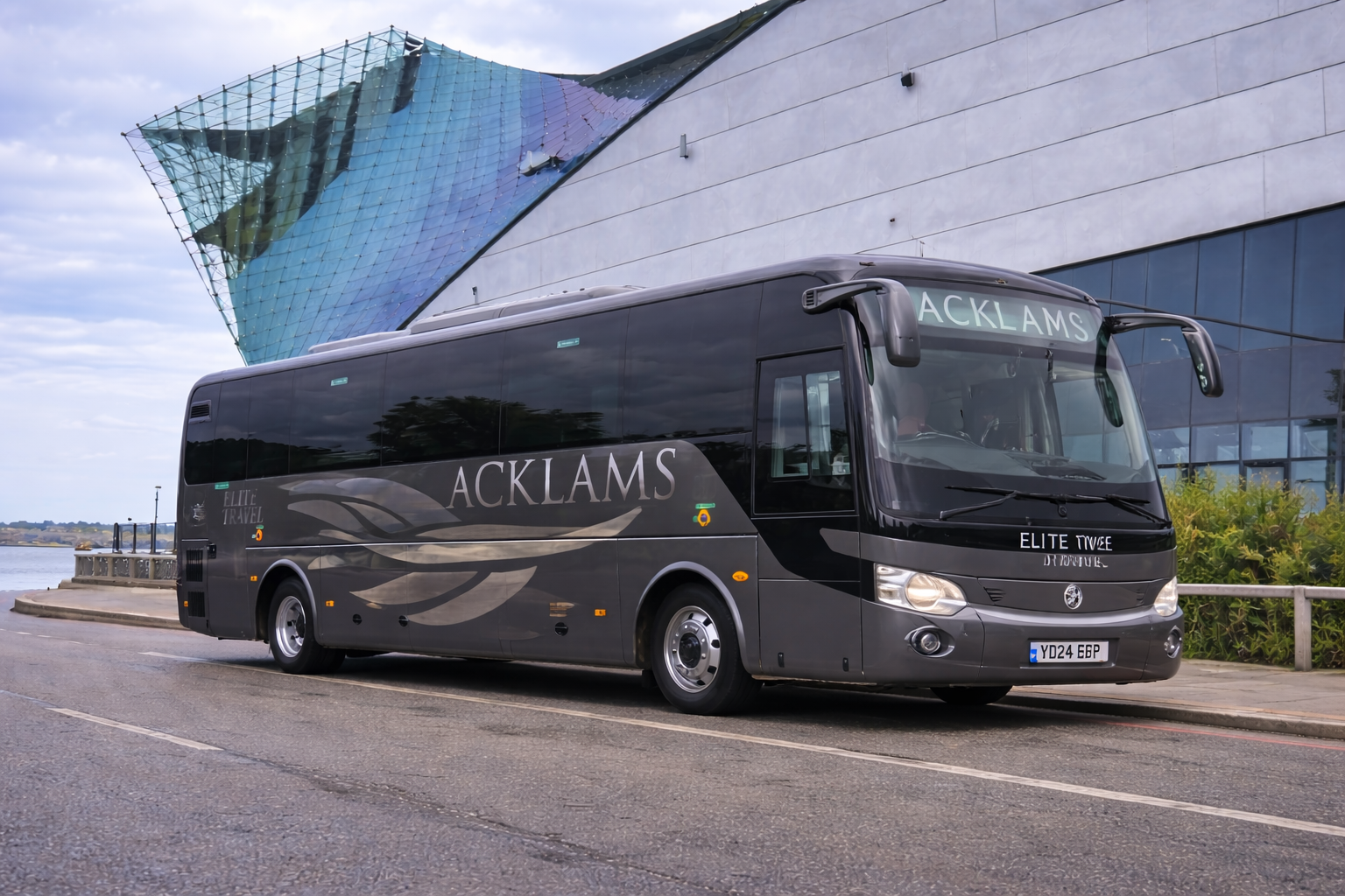 A black tour bus with the words 'ACKLAMS' and 'Elite Travel' parked on a street by a waterfront, with a modern building featuring colorful, iridescent glass panels in the background.