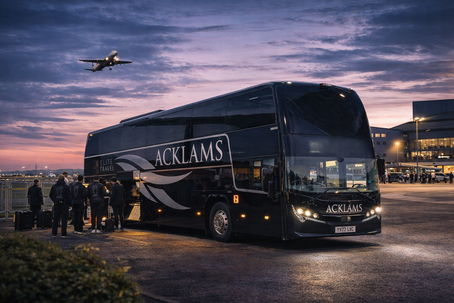 A black luxury coach bus with 'ACKLAMS' and 'Elite Travel' written on the side, parked at an airport during sunset. Several people are standing nearby with luggage, and an airplane is flying overhead in the colorful sky.