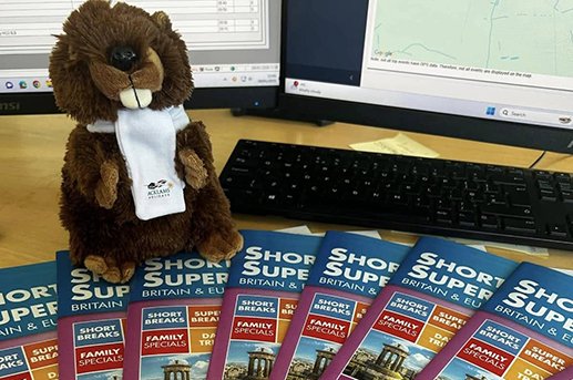 A plush beaver wearing a white tie sitting in front of computer monitors on a desk, with brochures about short breaks in Britain and Europe spread out in front.