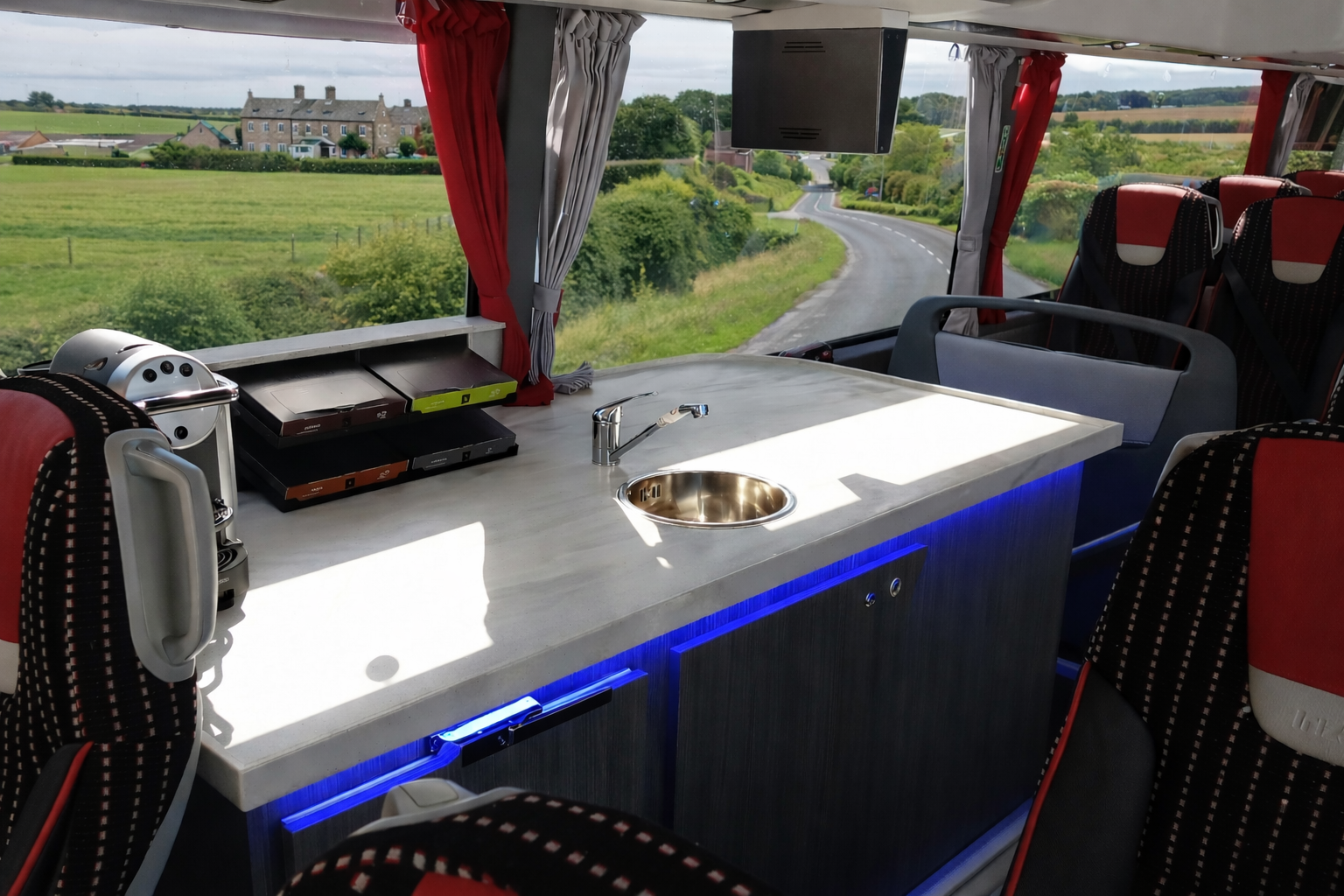 Interior of a bus with a white countertop, small sink, and built-in appliances, overlooking a rural landscape through the windows.